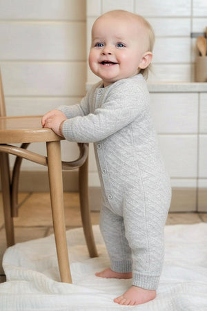 A smiling baby stands beside a wooden chair, wearing a light gray organic cotton jacquard romper with a diamond pattern. The setting features a cozy, neutral-toned kitchen environment.