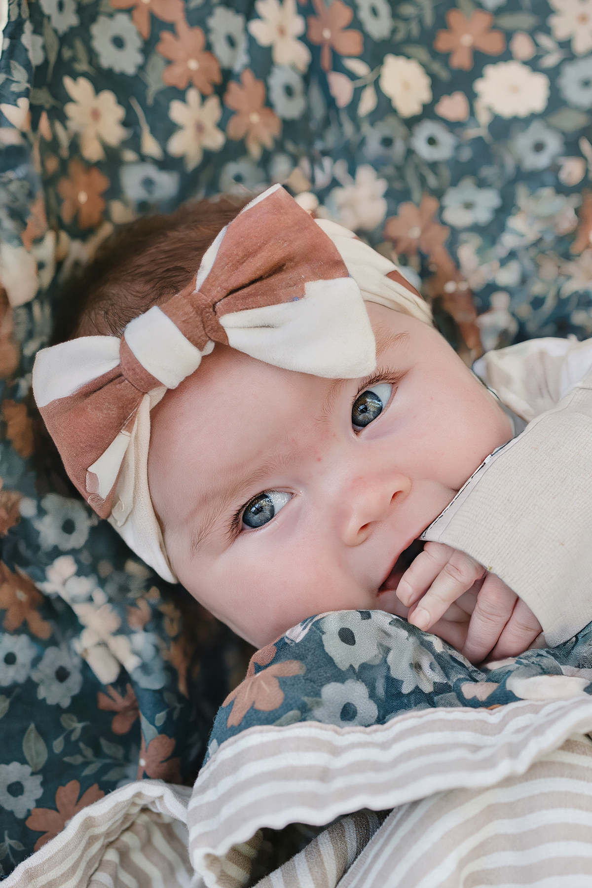 A close-up of a baby with bright blue eyes, wearing a rust checkered head wrap against a floral backdrop.