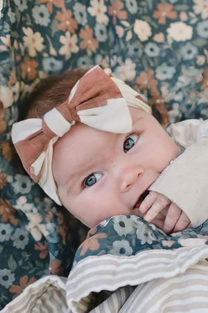 A close-up of a baby with bright blue eyes, wearing a rust checkered head wrap against a floral backdrop.