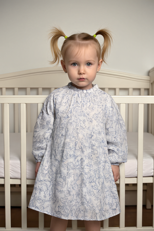 A young child stands in front of a crib, wearing a white dress with a delicate blue floral print and.