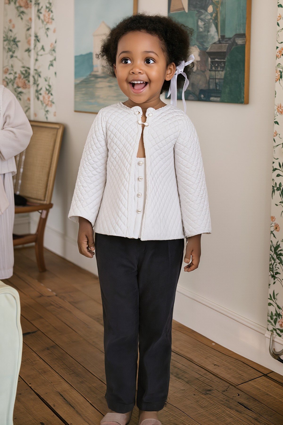 A smiling young girl wears a white quilted jacket and dark pants, standing in a cozy, decorated room.
