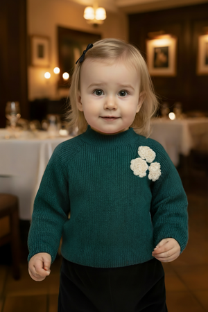 A toddler girl smiles while wearing a green mock neck sweater adorned with white rosettes, standing in a warmly lit.