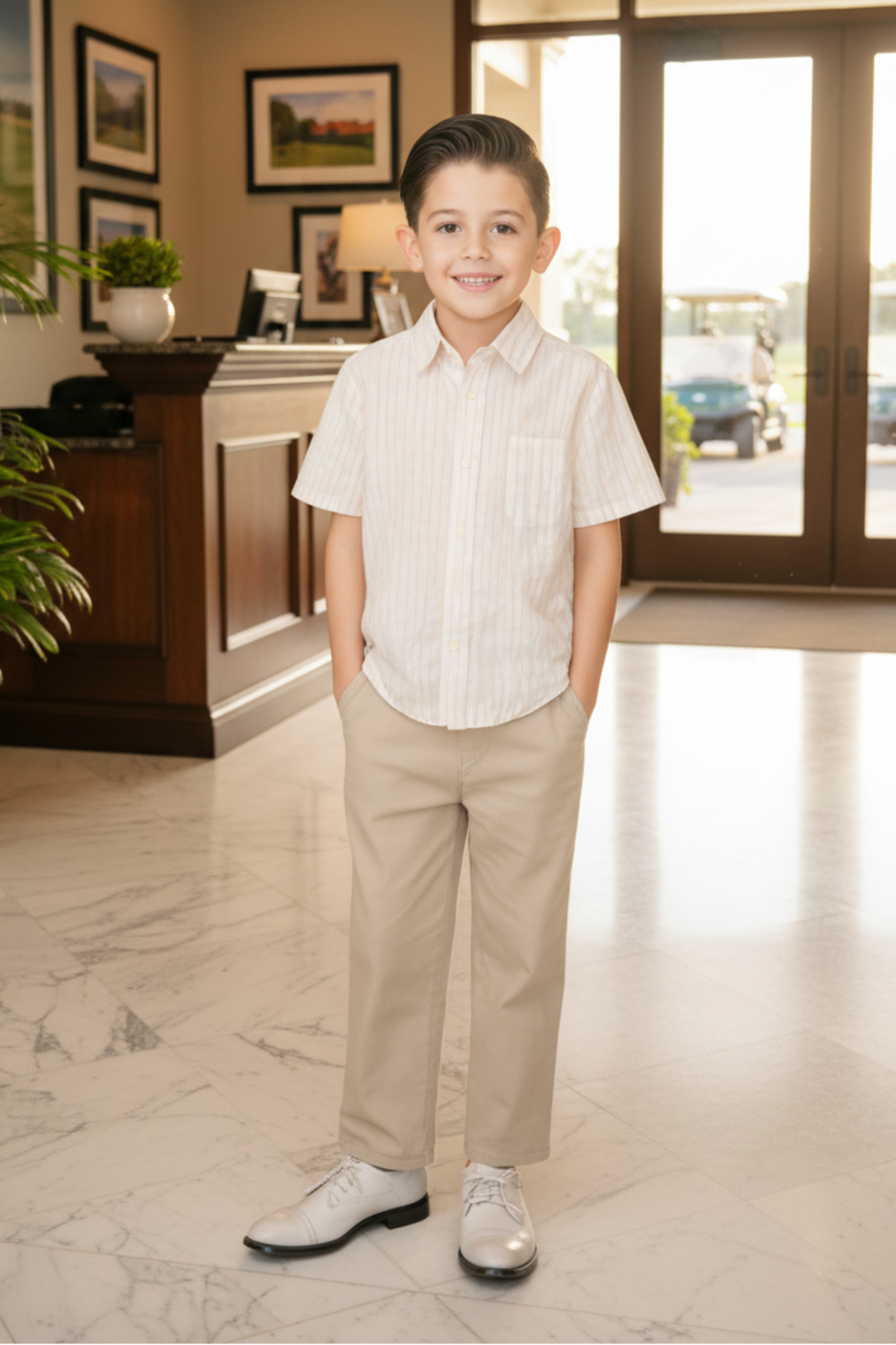 A smiling boy stands in a bright indoor setting, wearing a striped short-sleeve shirt and beige pants, with polished shoes.