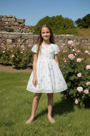 A girl stands in a garden wearing a white puff sleeve dress adorned with blue floral patterns, surrounded by pink.