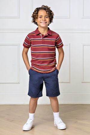 A smiling boy stands confidently in a maroon striped polo shirt and navy shorts, wearing white sneakers and socks.