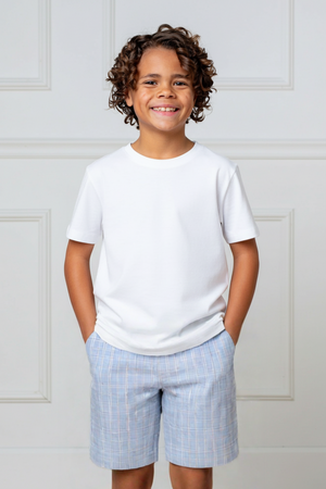 A smiling boy stands with hands in pockets, wearing a white t-shirt and blue checkered shorts against a neutral background.