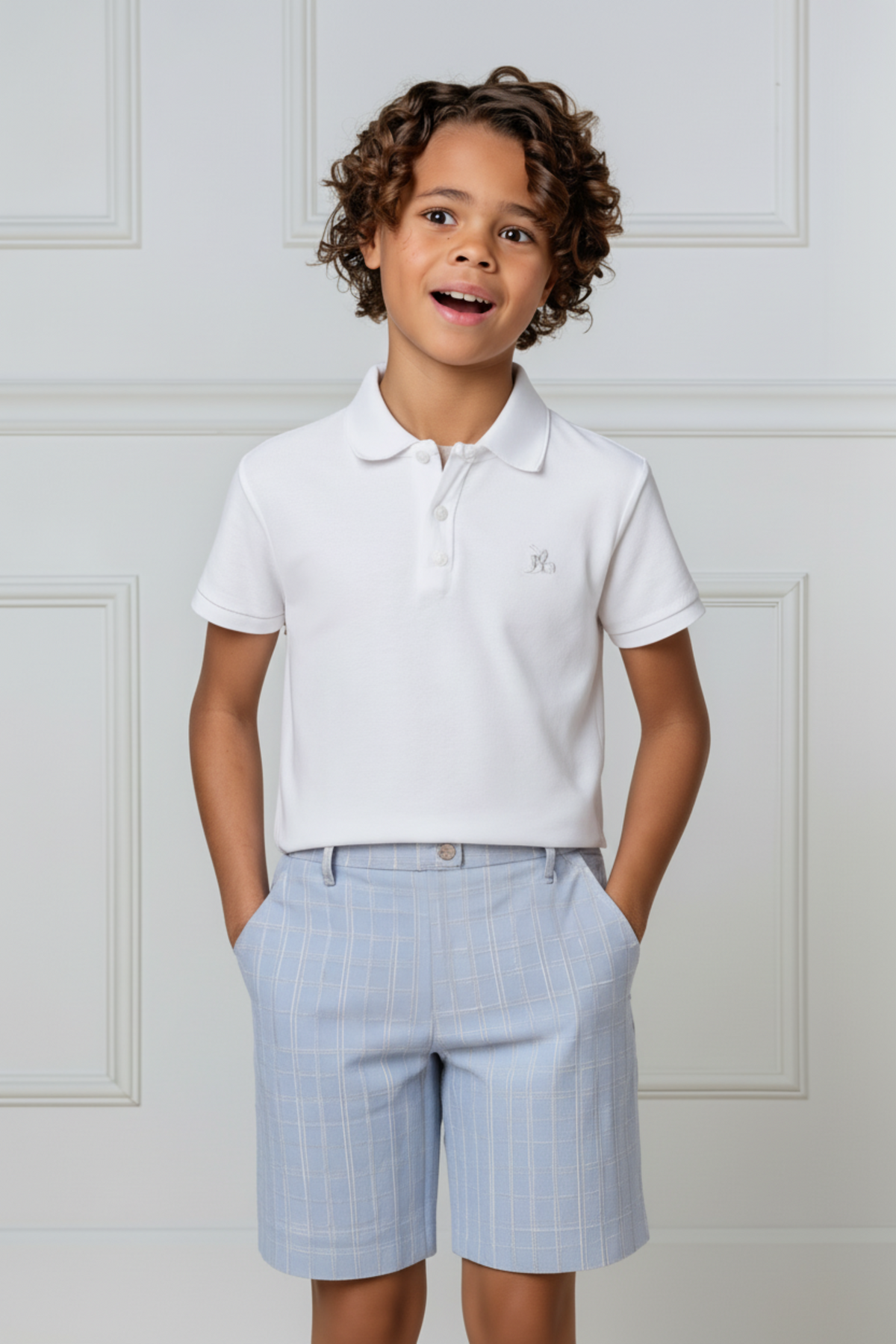 A young boy wears a white organic cotton polo shirt paired with light blue checkered shorts, smiling against a neutral.