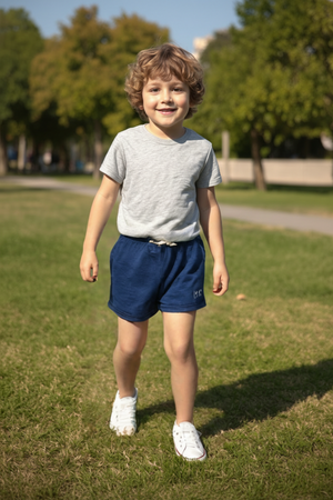 A smiling young boy with curly hair walks on a grassy path, wearing a gray t-shirt and navy cotton shorts. The outfit is comfortable and suitable for outdoor play.