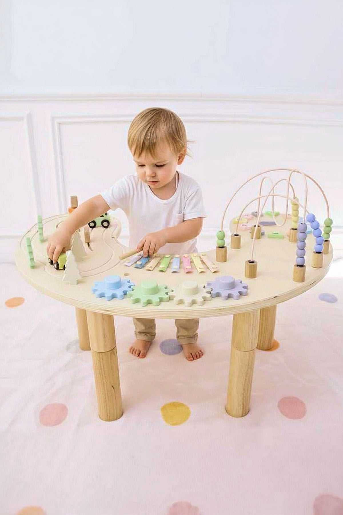 A child plays with a wooden multi-activity table featuring colorful gears, a xylophone, and a mini roadway, on a soft.