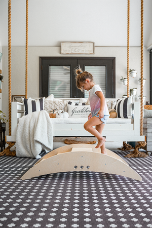 A young girl balances on a wooden climbing bridge inside a cozy living room, featuring a swing and decorative pillows. The natural wood adds warmth to the playful space.
