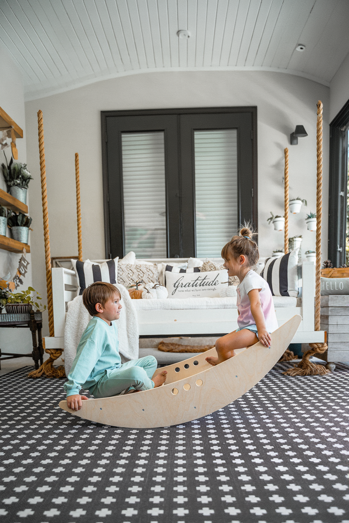 Two children are seated on a wooden rocking boat, smiling and playing together. The light-filled room features a cozy sofa and stylish decor, creating a welcoming atmosphere for active play.