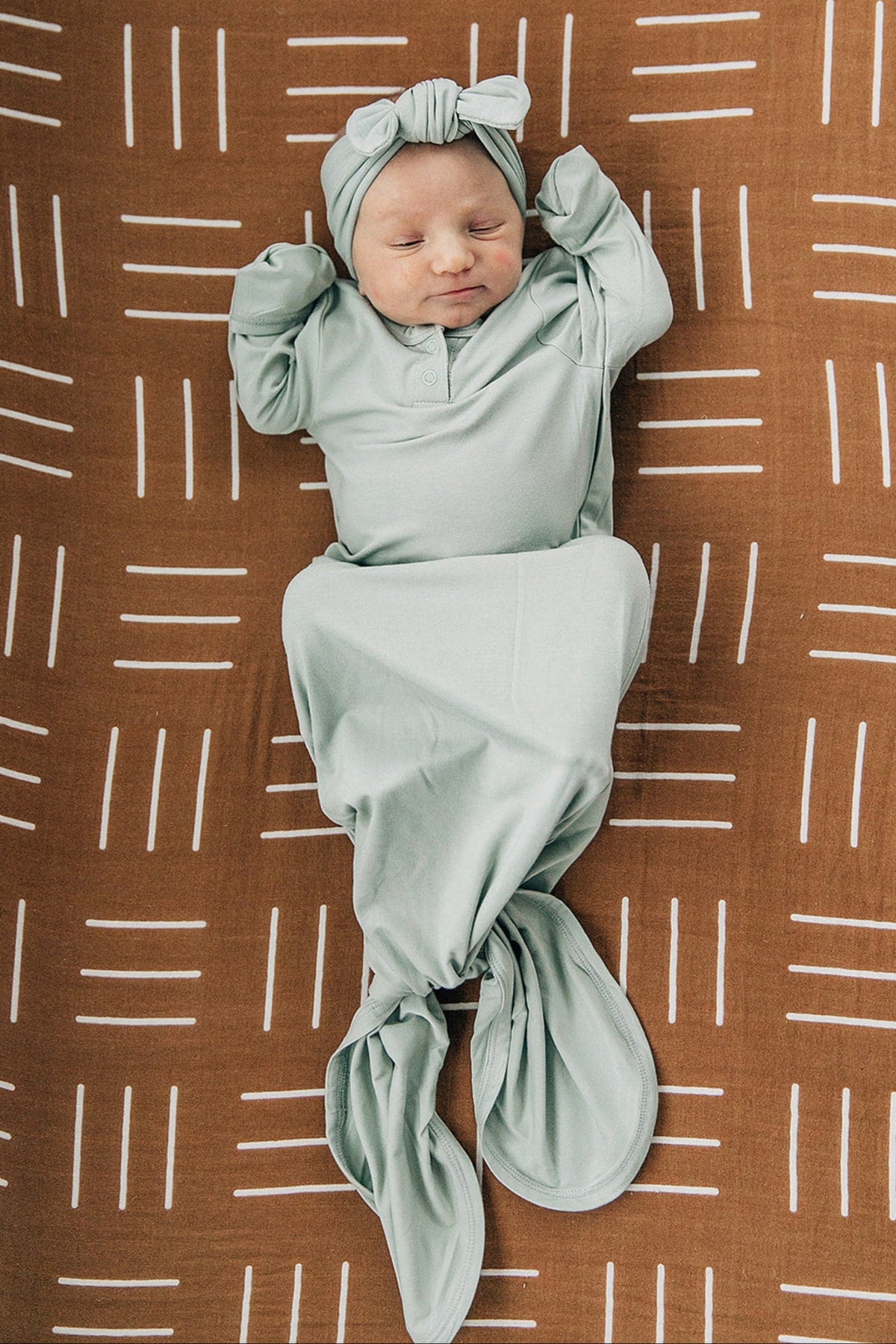 A baby in a sage green knot gown and headband lies on a brown patterned blanket.