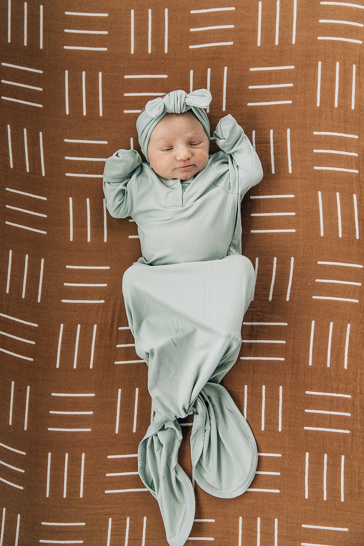 A baby in a light green outfit and headband lies on a mustard mudcloth-patterned crib sheet.