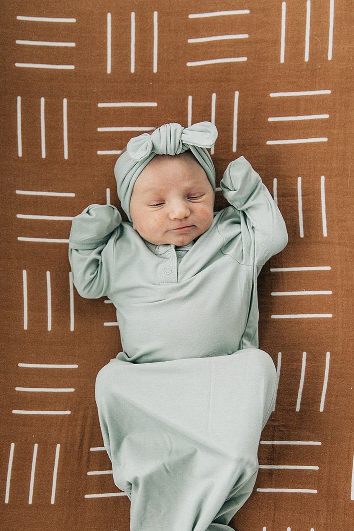 A baby in a sage bamboo head wrap and matching outfit lies on a patterned brown blanket.