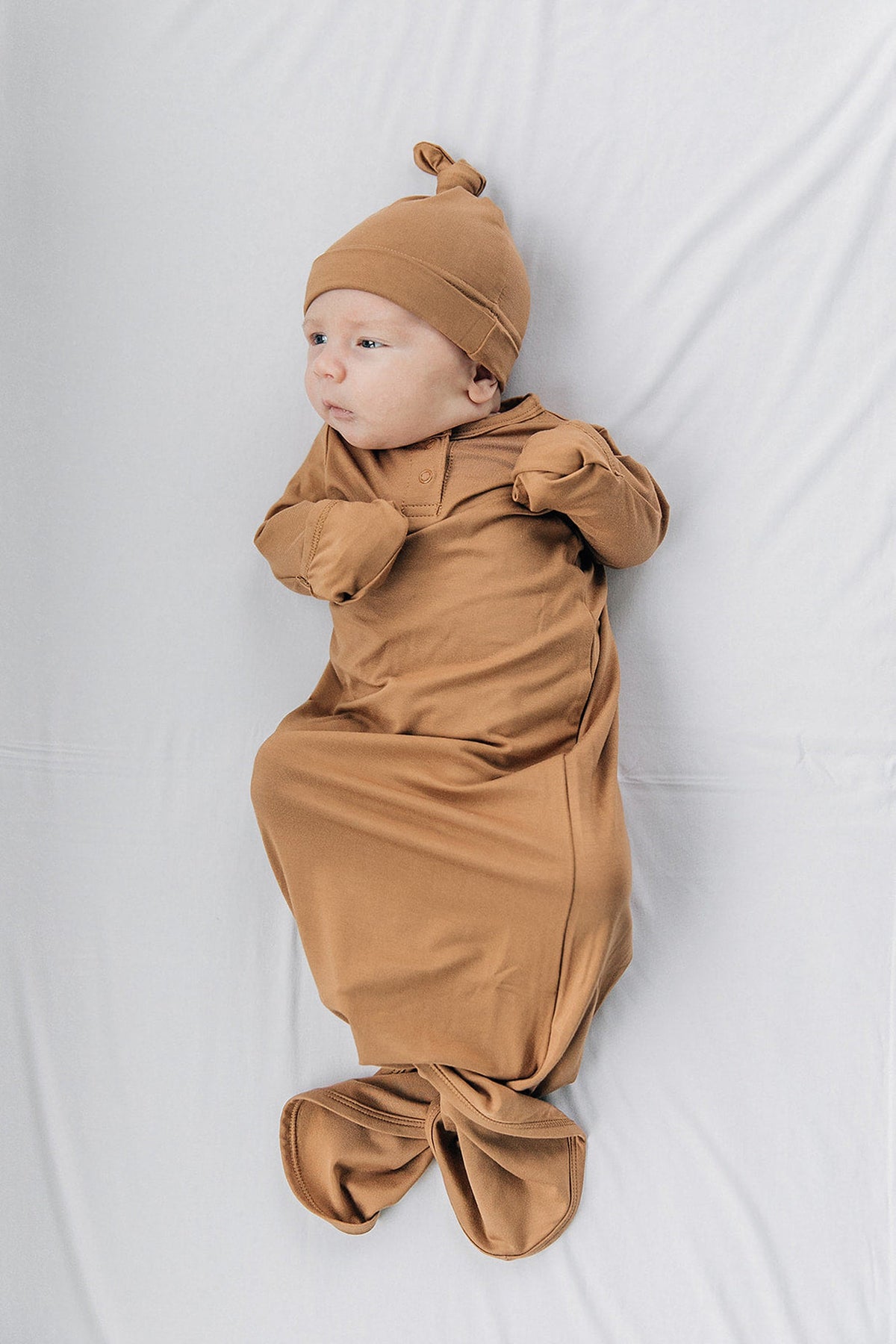 A baby wearing a mustard-colored knot gown and matching hat lies on a soft, light-colored surface.