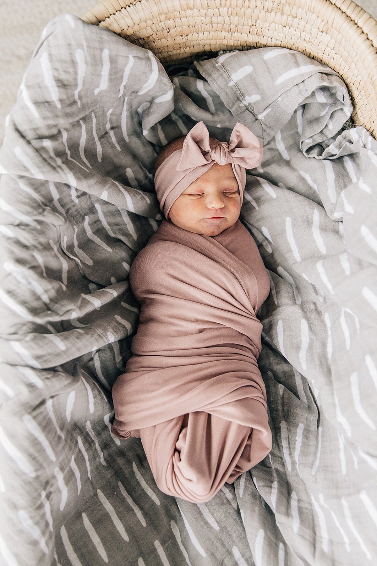 A baby is swaddled in a dusty rose bamboo blanket with a matching head wrap, lying on a patterned blanket.