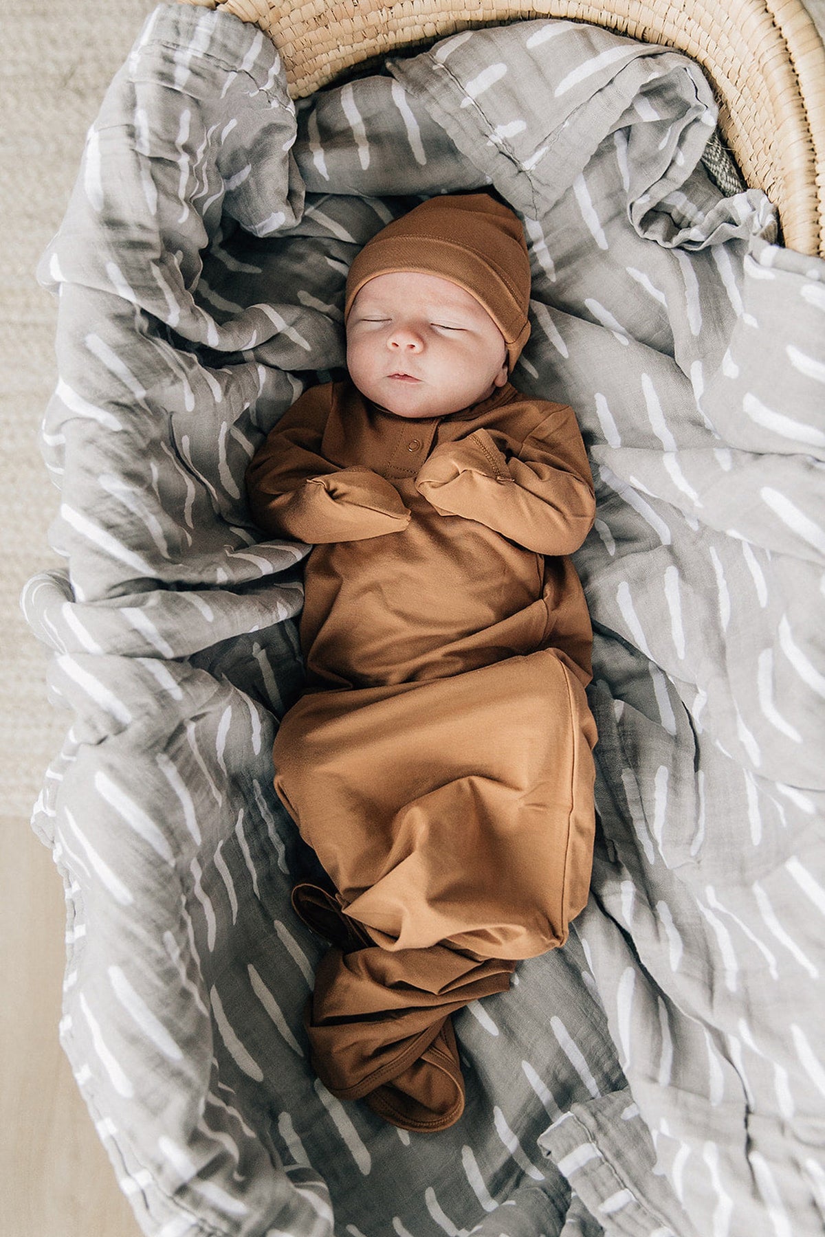 A baby wearing a mustard-colored gown and hat lies in a gray patterned blanket-lined basket.
