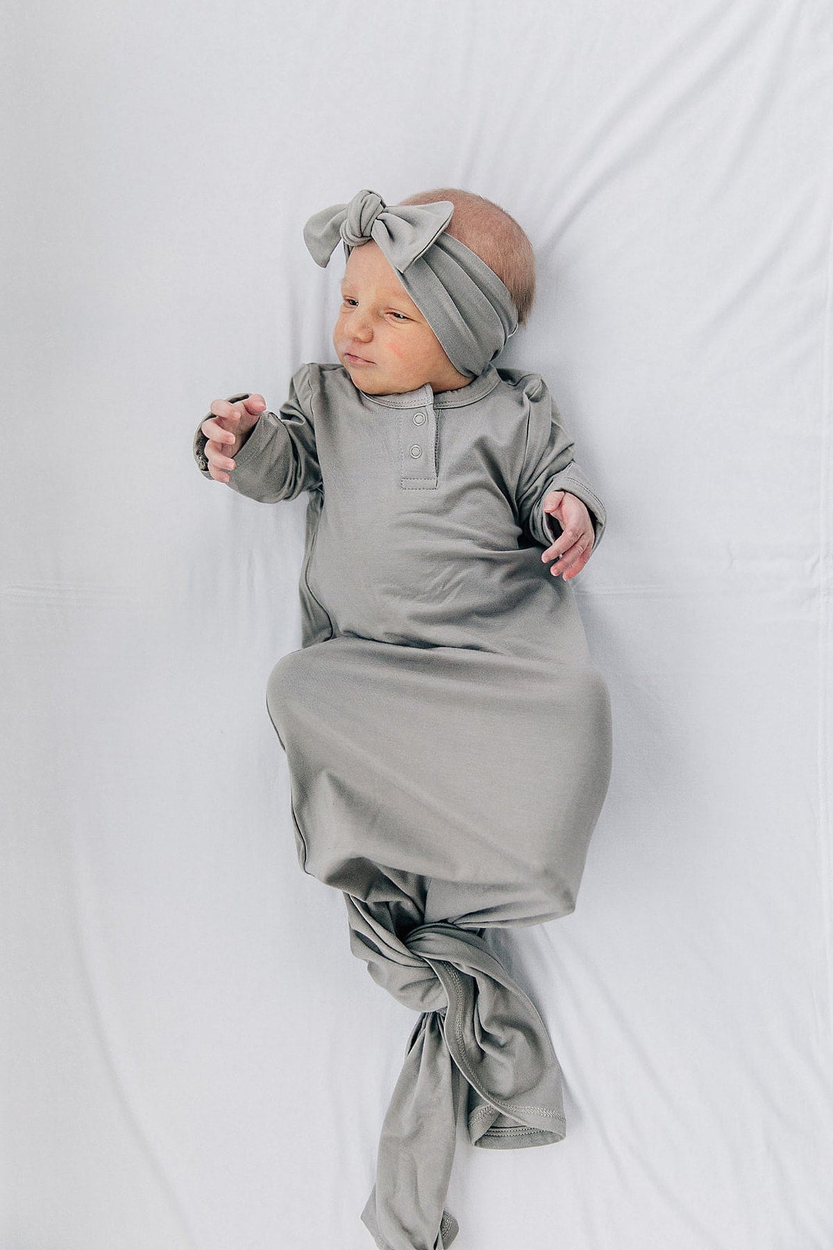 A baby wearing a light gray knot gown and headband lies on a white sheet, appearing comfortable and serene.