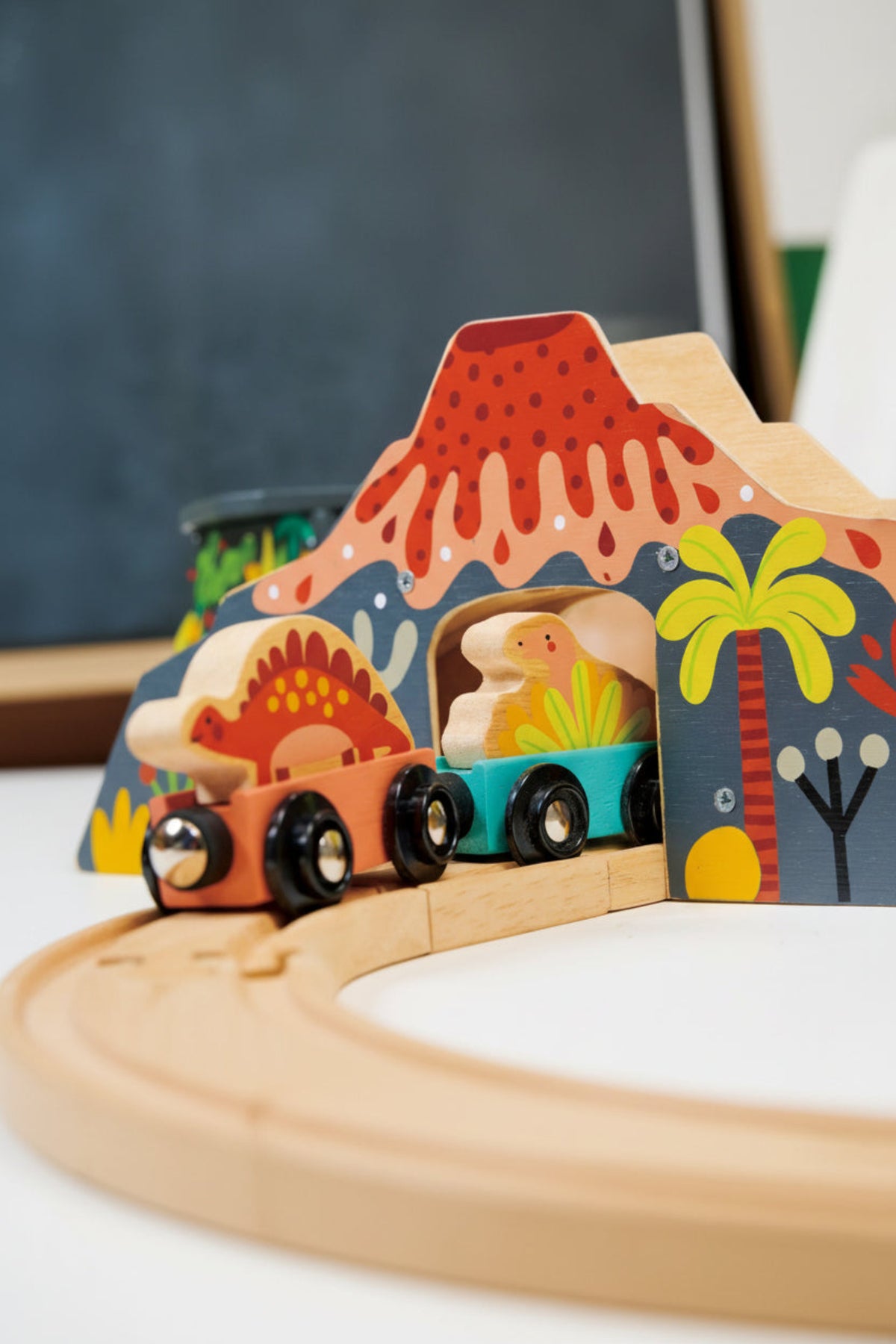 Wooden dinosaur-themed train set with colorful volcano and palm tree backdrop on a wooden track.