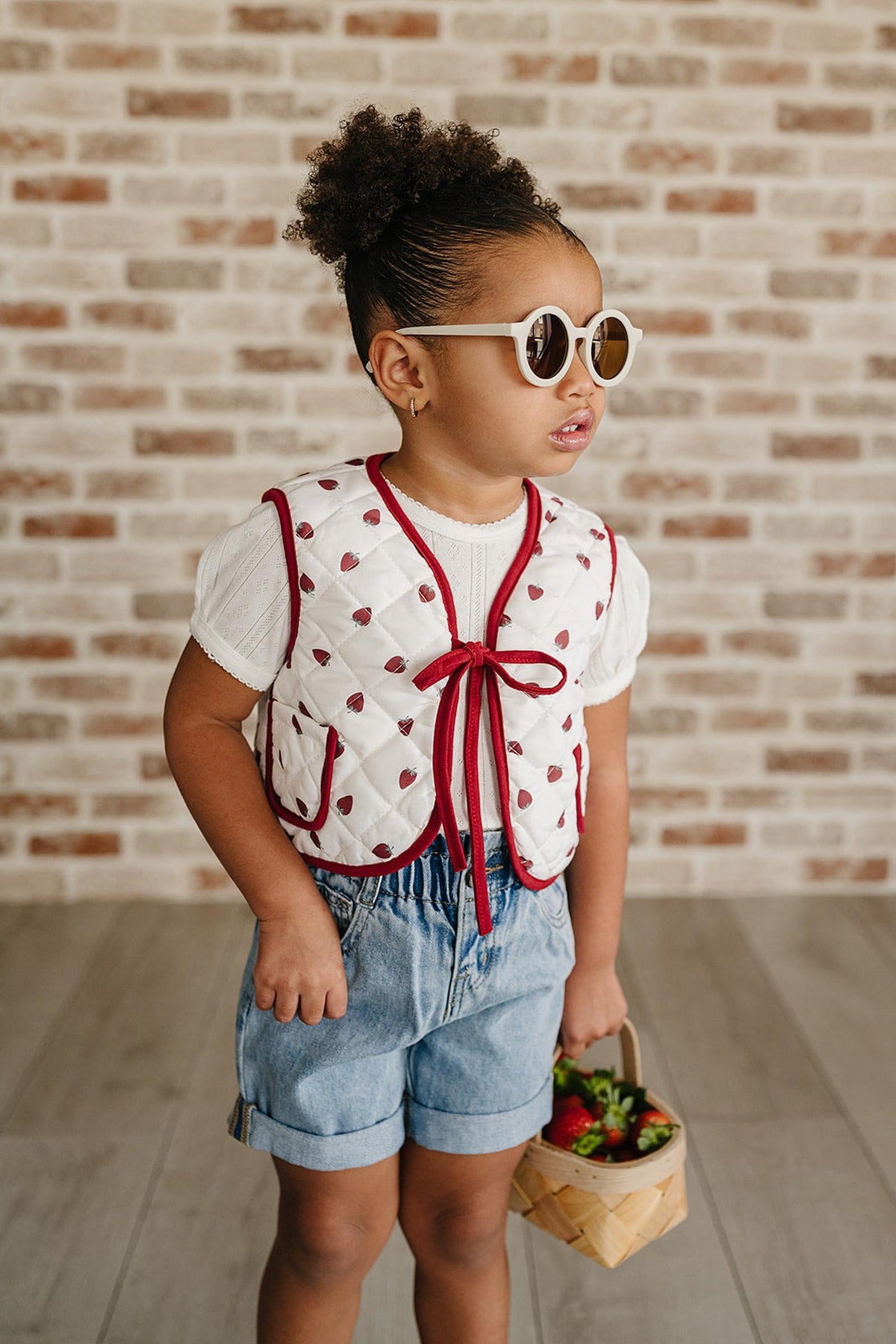 A young child poses in a strawberry-print quilted cotton vest with a red trim, paired with denim shorts and sunglasses.