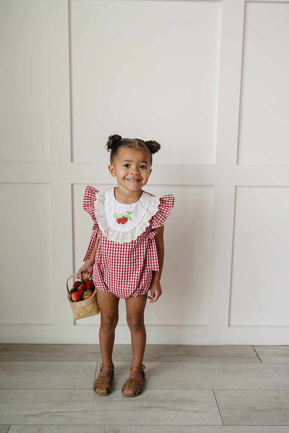 A young girl stands smiling, wearing a red gingham bubble romper with ruffle sleeves and a strawberry design. She holds a small basket of strawberries, set against a neutral backdrop.