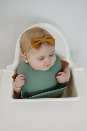 A baby in a high chair wearing a green silicone bib and a brown bow headband.