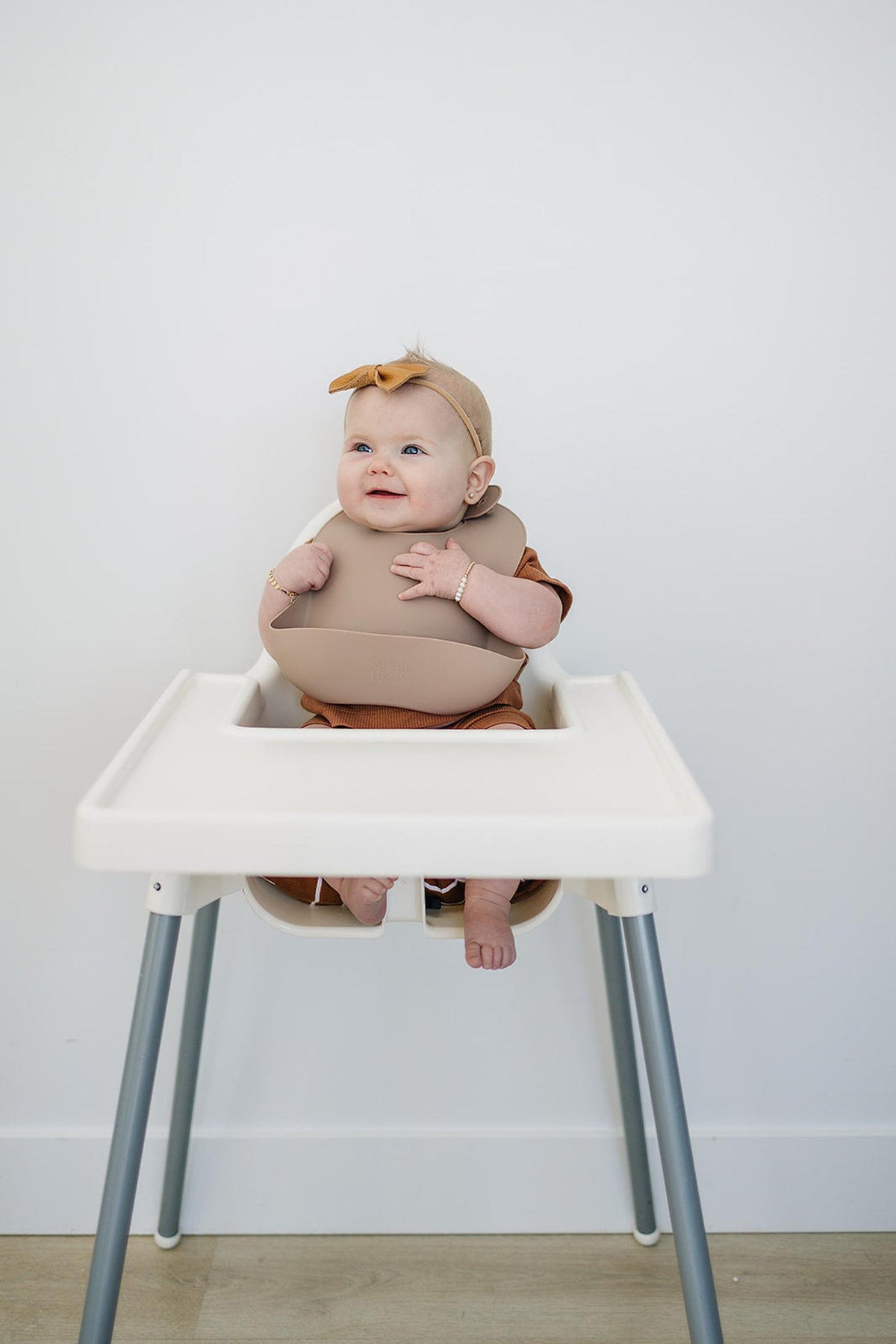 A baby wearing a tan silicone bib sits in a white high chair, smiling and wearing a matching headband.