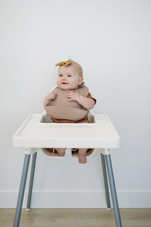 A baby wearing a tan silicone bib sits in a white high chair, smiling and wearing a matching headband.