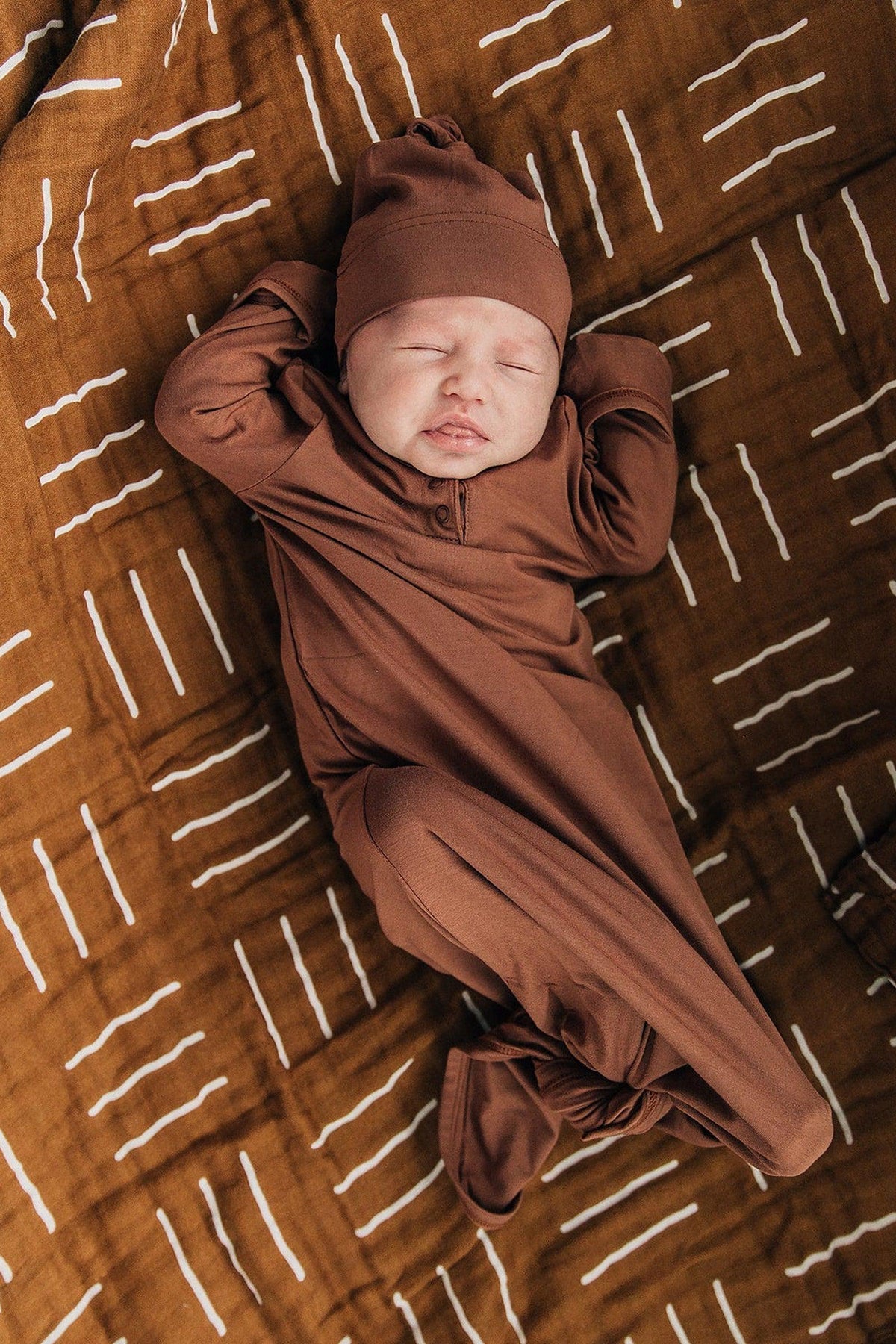 A baby in a brown outfit lies on a mustard mudcloth patterned blanket with white stripes.