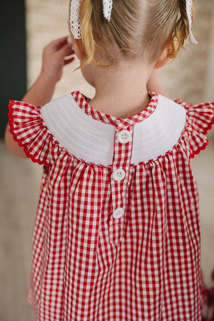 A child is seen from behind, wearing a red and white gingham dress with a white collar and decorative buttons.