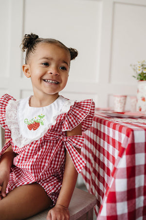 A smiling child sits at a table set with a red and white checkered tablecloth, wearing a red gingham bubble romper featuring strawberry accents and ruffle sleeves.