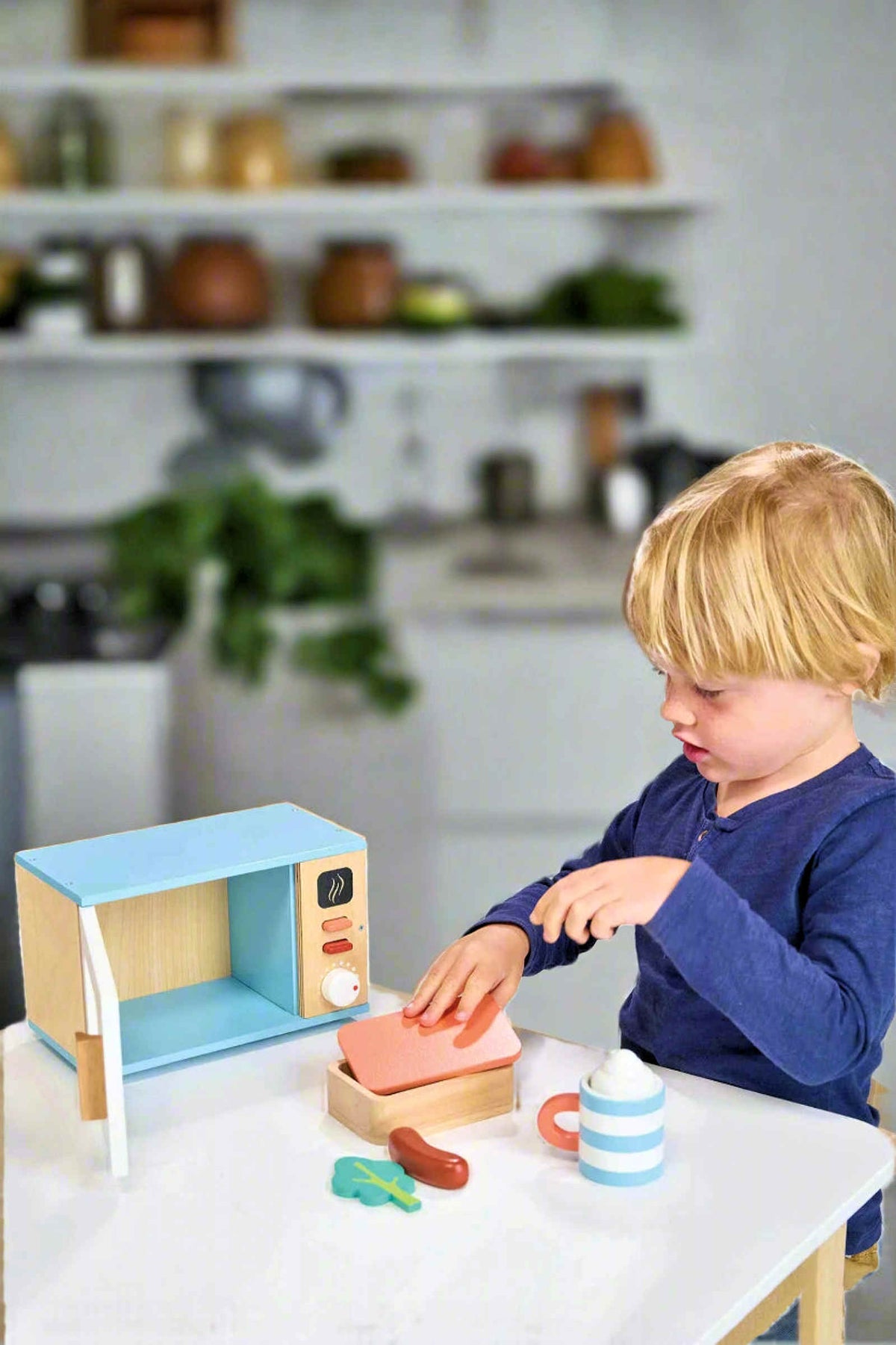 A child plays with a wooden toy microwave set, including a cup, sausage, broccoli, and wooden box on a table.