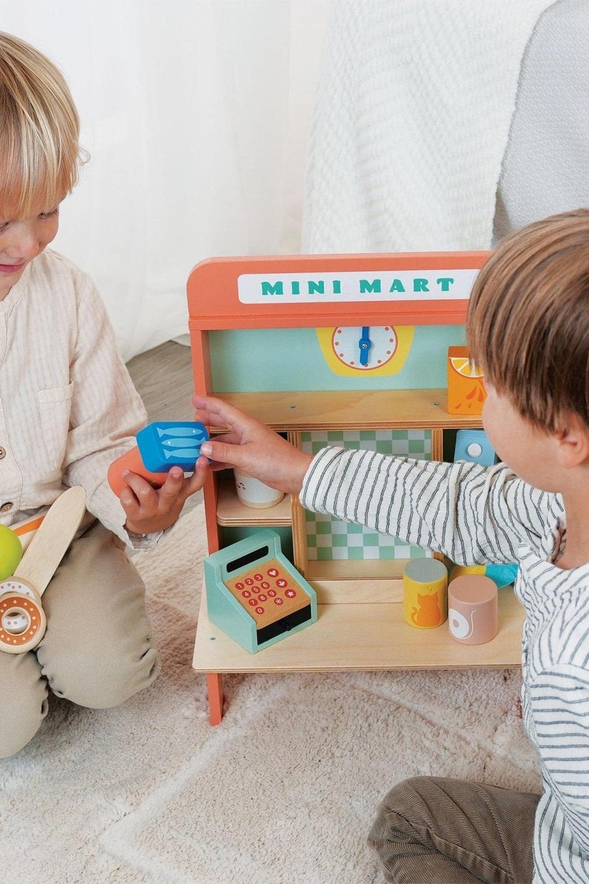 Two children play with a colorful wooden mini mart, arranging toy items and interacting with a cash register.