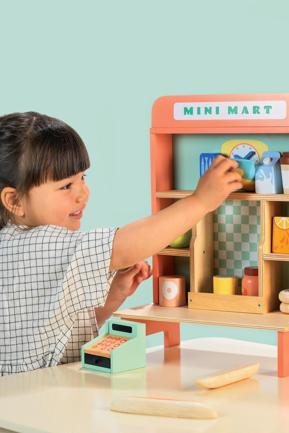 A child interacts with a colorful wooden Mini Mart playset, arranging toy food items on shelves and a cash register.