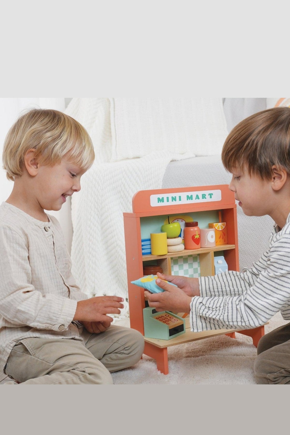 Two children play with a colorful wooden mini mart toy, exchanging items and smiling, surrounded by a cozy indoor setting.