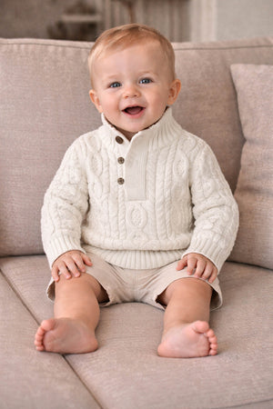 A smiling baby boy sits on a beige sofa, wearing a white cable-knit sweater with a mock neck and faux leather buttons, paired with light shorts.