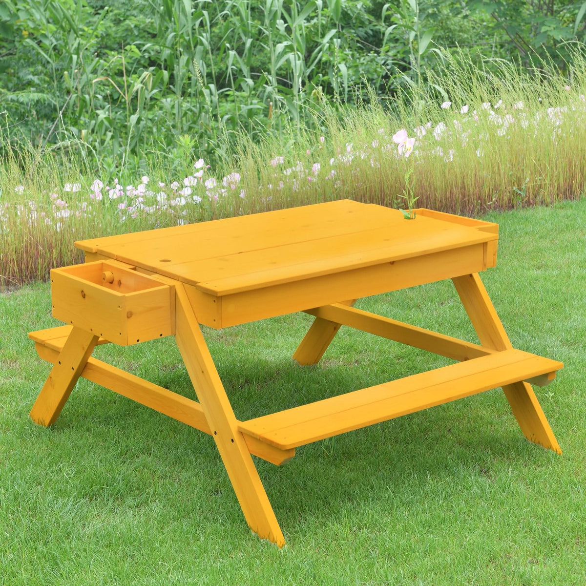 A vibrant yellow wooden picnic table with a liftable top and built-in drawer sits on green grass, surrounded by tall plants and flowers, perfect for outdoor play and activities.
