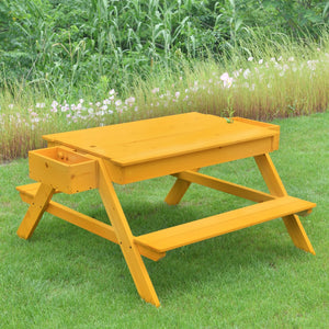 A vibrant yellow wooden picnic table with a liftable top and built-in drawer sits on green grass, surrounded by tall plants and flowers, perfect for outdoor play and activities.