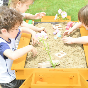 Two children play in a vibrant wooden sand table filled with light sand, surrounded by colorful toys and flowers, enjoying outdoor play in a grassy setting.