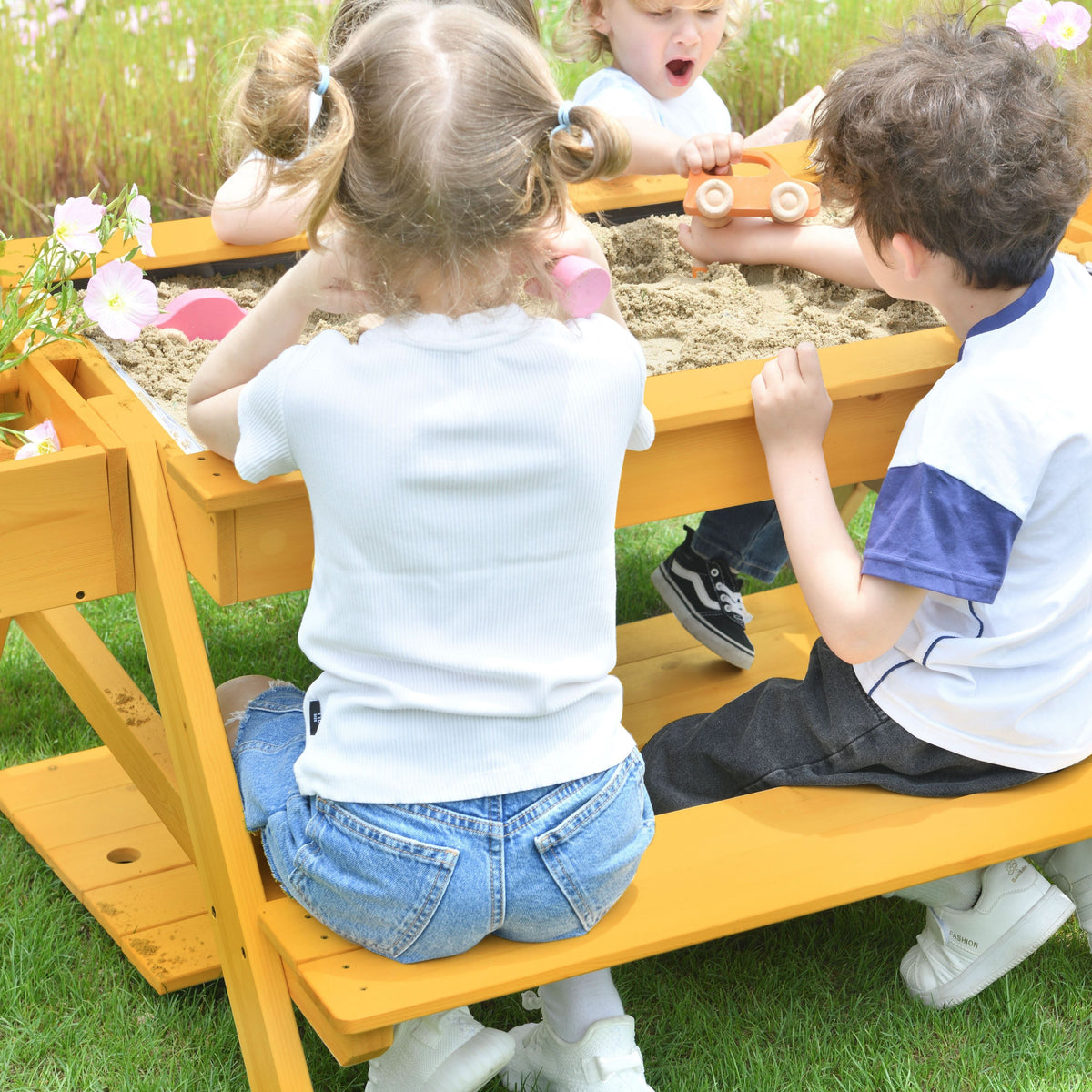 Three children play at a vibrant yellow picnic table, engaged in a sand area filled with sand toys. The setting features green grass and blooming flowers, emphasizing outdoor fun.