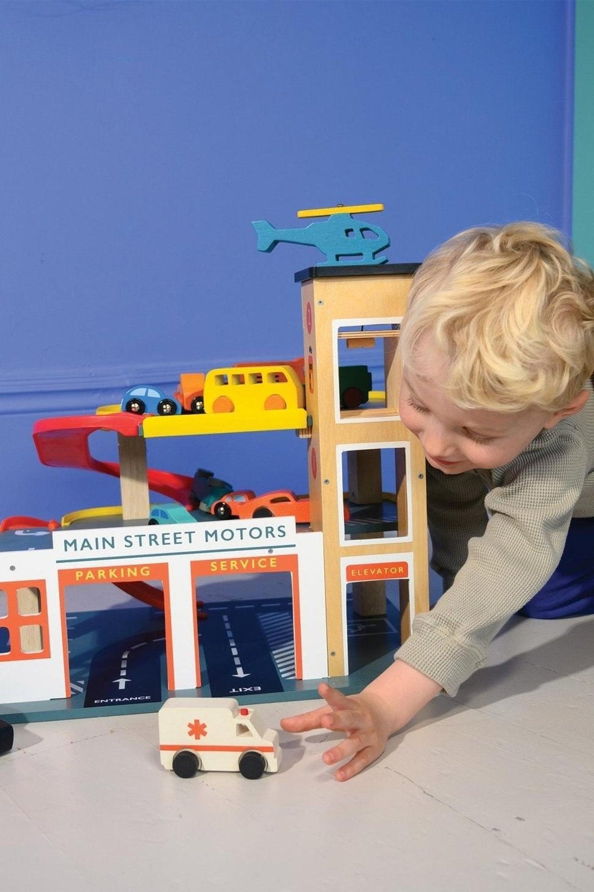 A child plays with a colorful wooden toy garage featuring ramps, cars, and a helicopter on top.