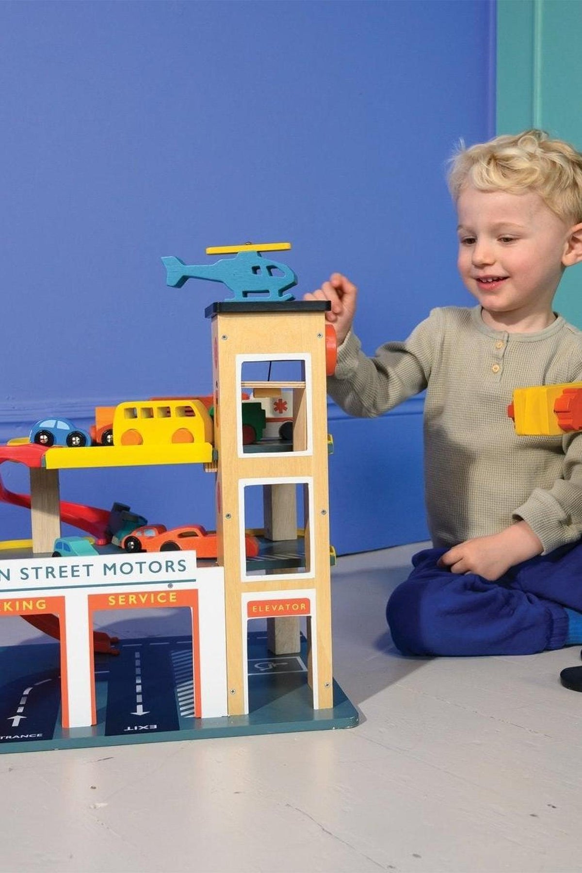 Child playing with a colorful wooden toy garage featuring cars, a helicopter, and ramps on a blue background.