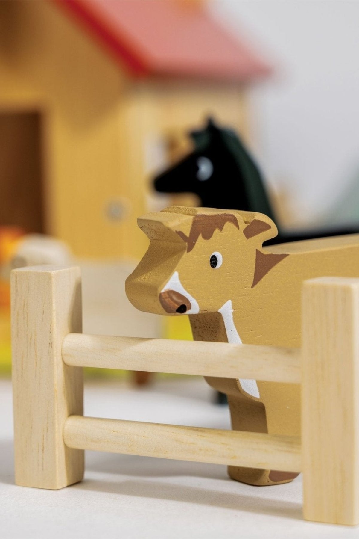 A wooden cow figure with brown and white markings stands near a wooden fence, set in front of a colorful barn and other farm elements, showcasing sustainable toy design for.