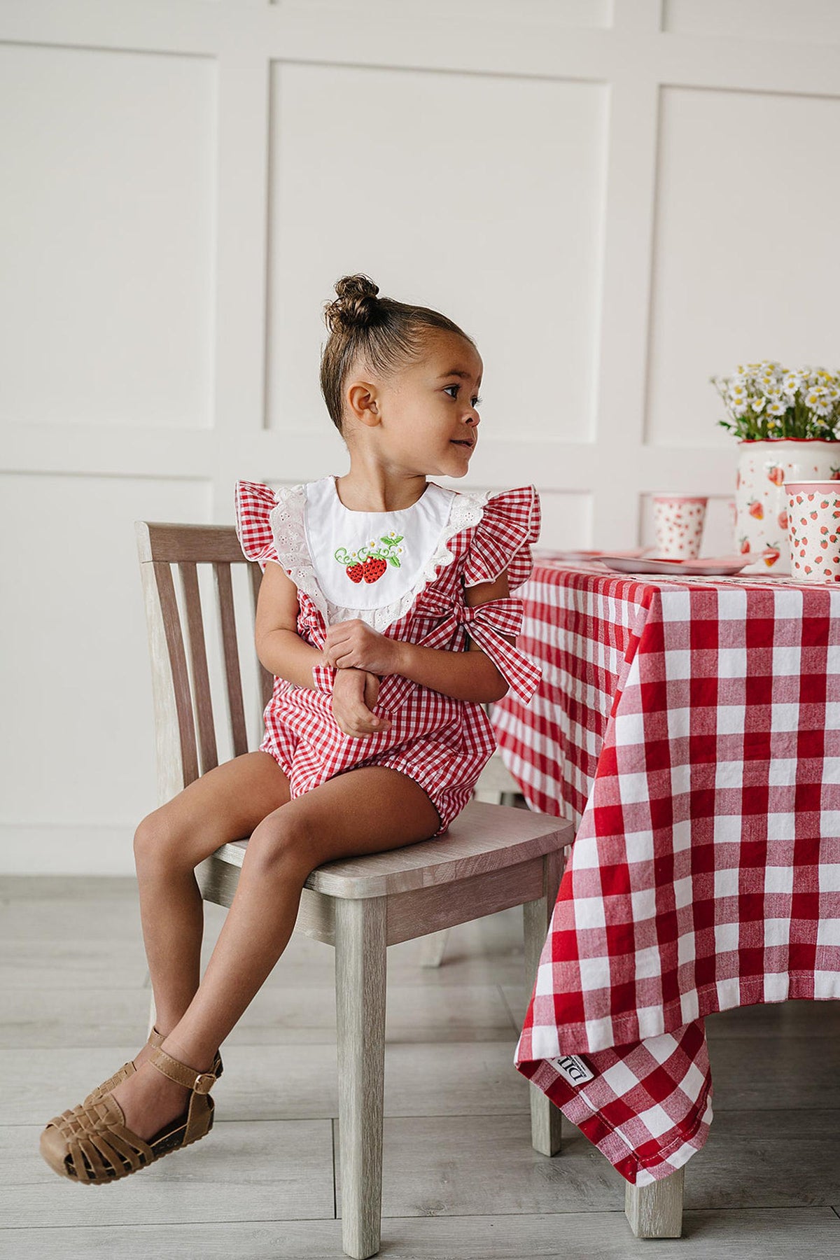 A young girl sits on a wooden chair wearing a red gingham bubble romper with ruffle sleeves and strawberry accents. The setting features a checkered tablecloth.