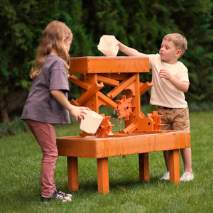 Two children play with a wooden water table in a lush green yard, pouring water through vibrant orange channels and gears, engaging in sensory exploration and imaginative play.