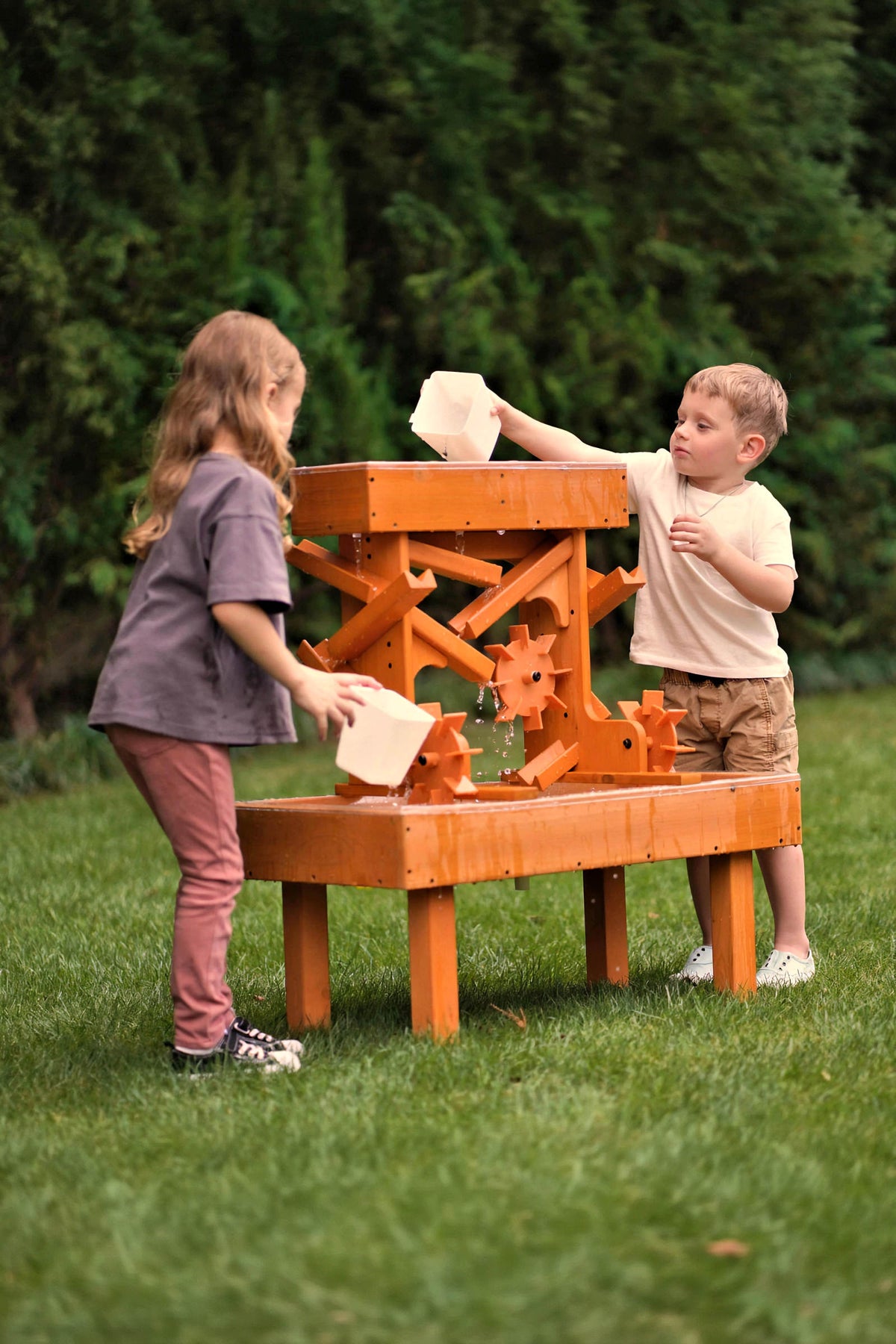 Two children play with an outdoor wooden water table, pouring water into the structures channels. The table features a bright orange finish and is set on a grassy area.