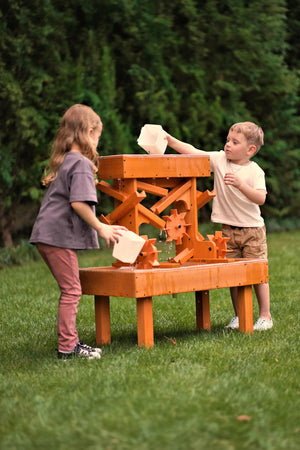 Two children play with an outdoor wooden water table, pouring water into the structures channels. The table features a bright orange finish and is set on a grassy area.