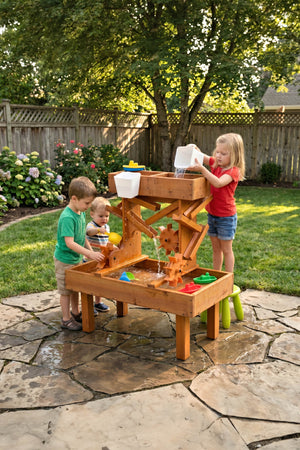 Two children play with an outdoor wooden water table in a sunny backyard, exploring water movement with colorful scoops and buckets.