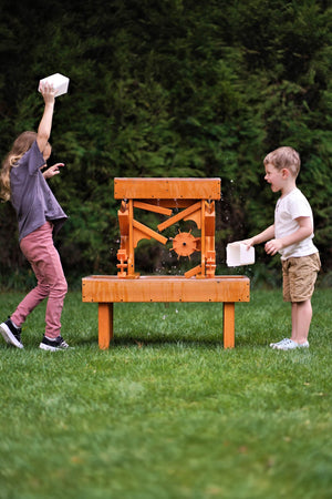 Two children play with a wooden water table outdoors, splashing water as they pour from white containers. The table features a vibrant orange finish, set against a green grassy backdrop.