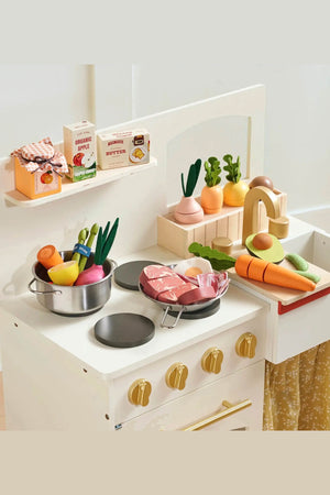 A wooden play kitchen features colorful, organic wooden play food, including vegetables and pretend meat, arranged on the countertop alongside food packaging. The kitchen is designed for imaginative play.